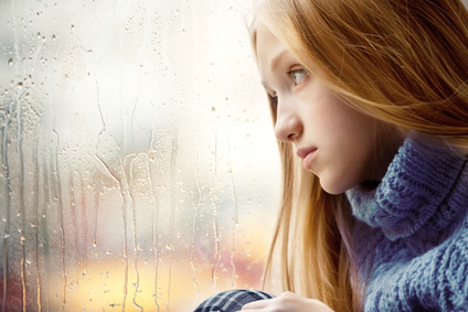 Emotional Portrait of a sad Girl with long blonde Hair. She is looking through a Window in the Rainy Autumn Day