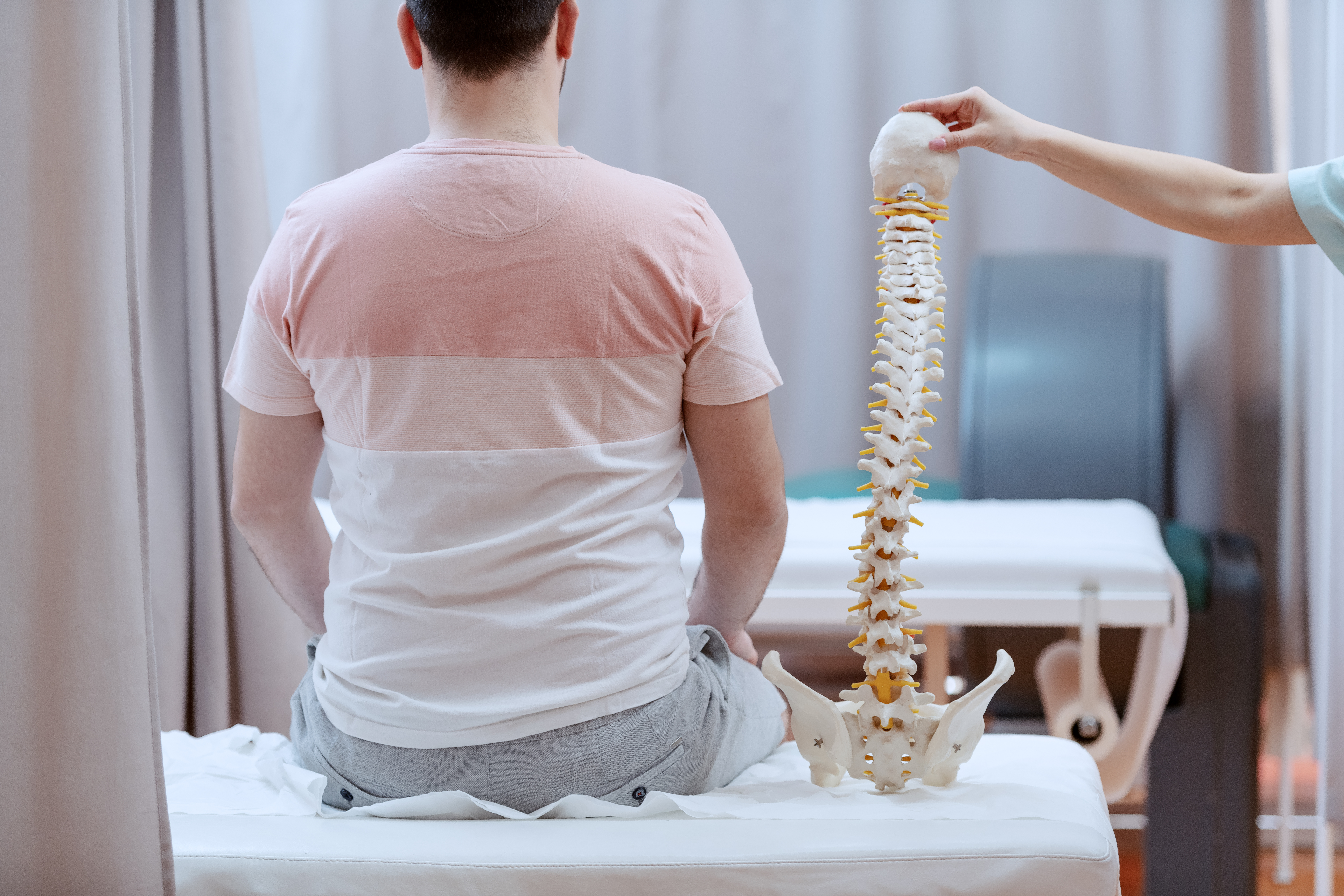 Male patient sitting on the hospital bed with backs turned. Next to him is nurse holding spine model.