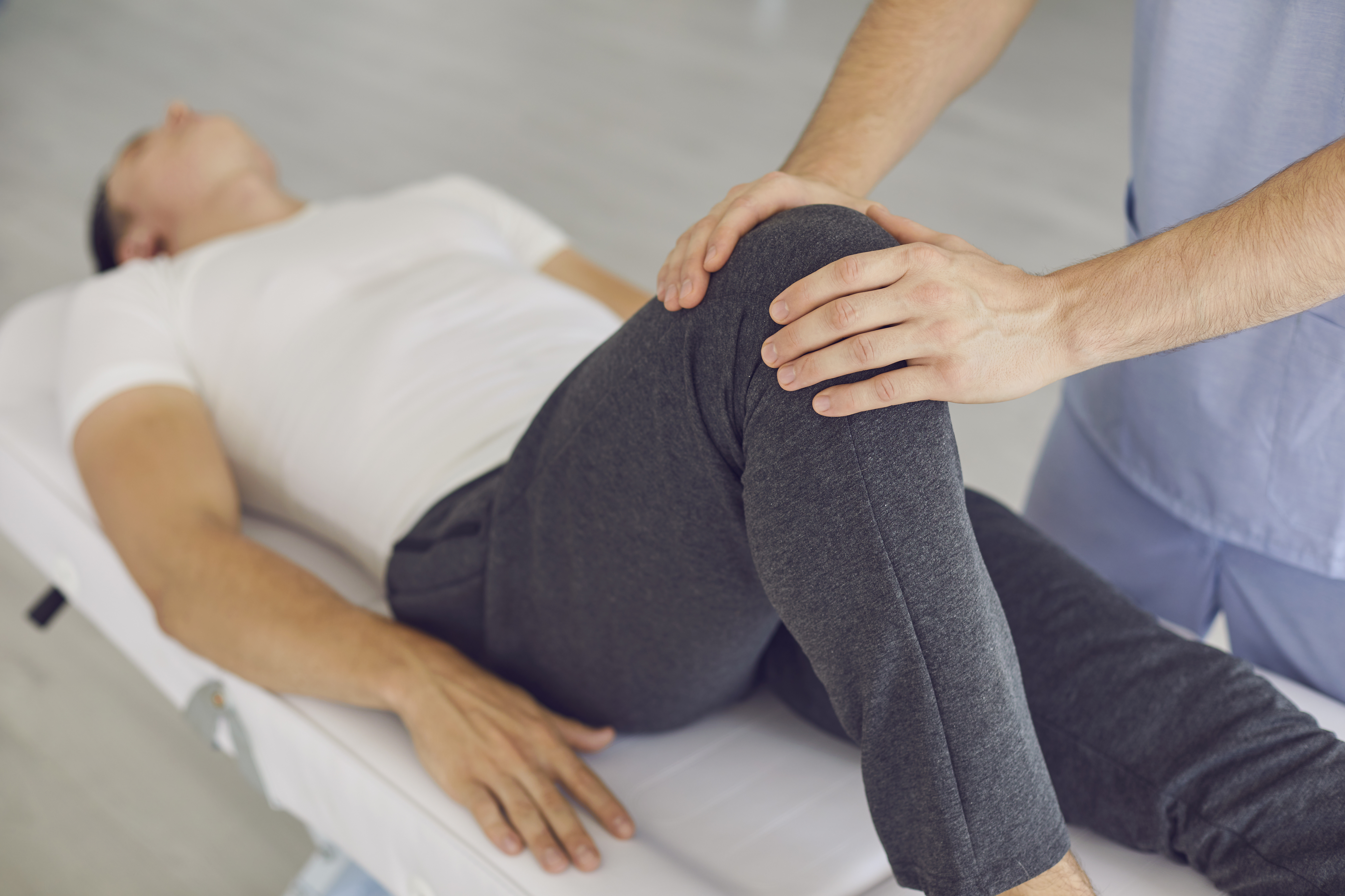 Hands of professional man chiropractor manual therapist fixing lying patients knee in stable position during visit at rehabilitation theapy in manual therapy clinic. Osteopath during work concept